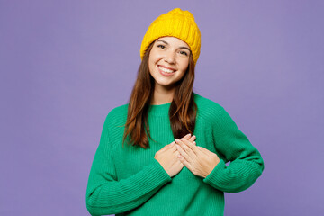 Sticker - Young smiling cheerful woman she wear green sweater yellow hat casual clothes put folded hands on heart look camera isolated on plain pastel light purple background studio portrait. Lifestyle concept.