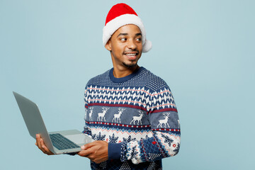 Wall Mural - Side view young IT man wear knitted sweater Santa hat posing hold use work on laptop pc computer look aside isolated on plain blue background Happy New Year 2024 celebration Christmas holiday concept