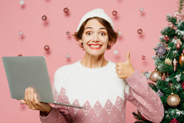 Wall Mural - Merry fun young IT woman wear white sweater hat posing hold use work on laptop pc computer show thumb up isolated on plain pastel pink background. Happy New Year celebration Christmas holiday concept.