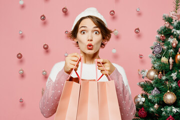 Canvas Print - Merry surprised young woman wear white sweater hat posing hold paper package bags after shopping isolated on plain pink background. Happy New Year celebration Christmas holiday sale buy day concept.