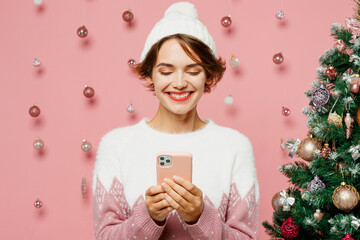 Canvas Print - Merry young woman wear white sweater hat posing hold in hand use mobile cell phone typing message isolated on plain pastel pink background studio. Happy New Year celebration Christmas holiday concept.