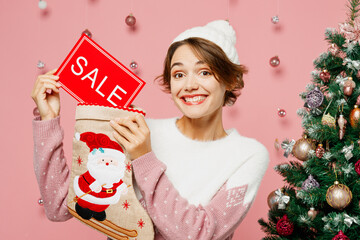 Wall Mural - Merry young woman wear white sweater hat posing hold in hand stocking card with sale title text isolated on plain pastel pink background studio. Happy New Year celebration Christmas holiday concept.