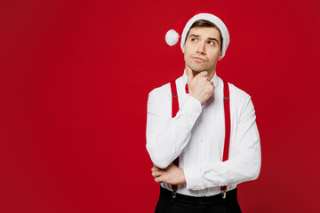 Wall Mural - Merry young man wear white shirt Santa hat posing put hand prop up on chin, lost in thought and conjectures isolated on plain red background studio Happy New Year Christmas celebration holiday concept