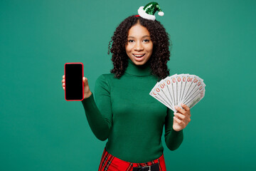 Wall Mural - Merry little kid teen girl wears hat casual clothes posing hold blank screen mobile cell phone, fan cash money in dollar banknotes isolated on plain green background. Happy New Year Christmas concept.