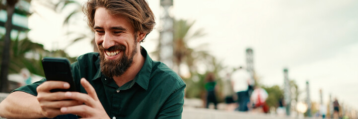 Wall Mural - Close-up portrait of a man with a beard recording a voice message on the embankment background. Closeup of a young hipster male using a mobile phone