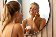 Beautiful smiling woman applying moisturizer cream on her face looking in mirror. Skin care, cosmetology, anti aging concept 