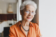 Closeup indoor portrait of elderly wise kind female grandmother in glasses with gray hair in orange shirt leaning against wall at home on blurred kitchen background, looking at camera with smile