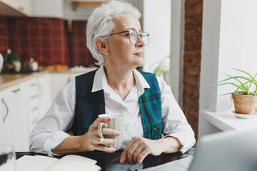 Wall Mural - Caucasian gray-haired elderly woman accountant in glasses and white shirt sitting at kitchen table with cup of hot tea, looking aside through window, having rest after working online