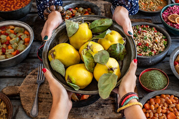 Wall Mural - Fresh and ripe quinces a the hand of two women in the copper bowl. Many types of vegan foods on the table with fruits. 