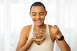 Smiling millennial latin lady in sportswear eat muesli cereal, enjoy breakfast