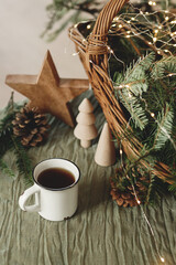 Wall Mural - Stylish cup of warm tea, fir branches in basket, wooden trees and star, pine cones and lights on table. Modern christmas rustic eco friendly decor, scandinavian winter hygge. Atmospheric christmas