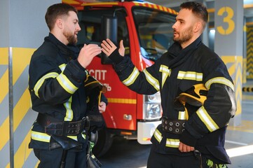 Wall Mural - Two firefighters in protective clothing in helmets with fire engine