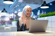 © maya1313 - Cheerful muslim lady in hijab working online at cafe, using laptop, copy space. Side view of smiling arab woman freelancer typing on laptop, enjoying her distant job, Muslim working woman concept.