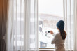 young woman in sweater with cup of coffee looking through the window in winter season, happy female enjoying snowfall outdoor view at apartment or home in the morning. Waking and Relaxing concept