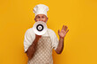 old grandfather chef in apron and hat announces information into megaphone on yellow isolated background