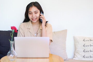 happy asian woman sitting on sofa answer call on smartphone and working with laptop wearing casual clothes, female designer working on project at home