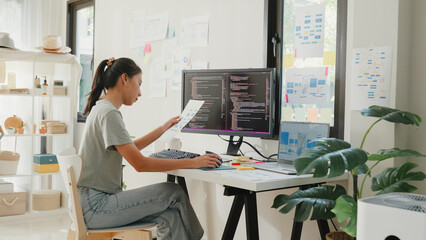 Side view of professional young Asia girl IT development programmer typing on keyboard coding programming fixing data code on computer screen and laptop on table in workroom at house office.