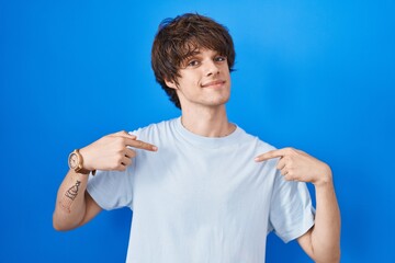 Sticker - Hispanic young man standing over blue background looking confident with smile on face, pointing oneself with fingers proud and happy.