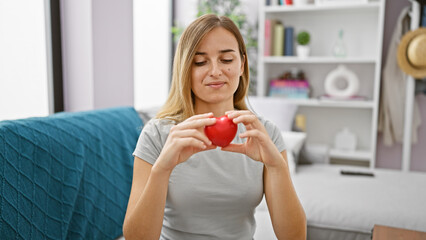 Poster - Confident young adult woman, enjoy the indoors lifestyle, sitting comfortably on her cozy living room sofa, holding a heart, with a big smile lighting up her beautiful, blonde portrait.