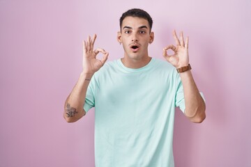 Wall Mural - Handsome hispanic man standing over pink background looking surprised and shocked doing ok approval symbol with fingers. crazy expression