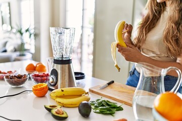 Young beautiful hispanic woman preparing vegetable smoothie with blender peeling banana at the kitchen