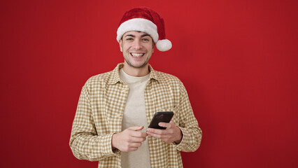 Canvas Print - Young hispanic man wearing christmas hat using smartphone over isolated red background