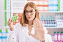 Young Redhead Woman Working At Pharmacy Drugstore Holding Birth Control Pills With Open Hand Doing Stop Sign With Serious And Confident Expression, Defense Gesture