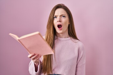 Canvas Print - Young caucasian woman reading a book over pink background angry and mad screaming frustrated and furious, shouting with anger looking up.