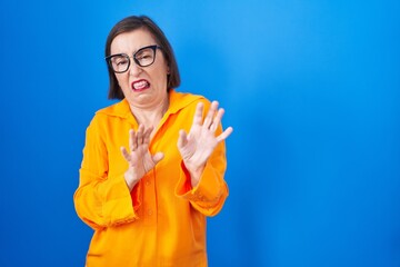 Wall Mural - Middle age hispanic woman wearing glasses standing over blue background disgusted expression, displeased and fearful doing disgust face because aversion reaction. with hands raised