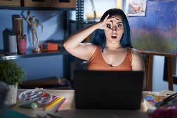 Wall Mural - Young modern girl with blue hair sitting at art studio with laptop at night doing ok gesture shocked with surprised face, eye looking through fingers. unbelieving expression.