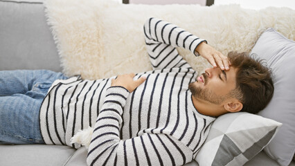 Canvas Print - Troubled young arab man enduring head and stomachache, lying unhappily at home, struggling with illness