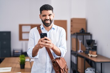 Canvas Print - Hispanic man with beard using smartphone at the office winking looking at the camera with sexy expression, cheerful and happy face.