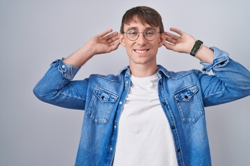 Poster - Caucasian blond man standing wearing glasses smiling pulling ears with fingers, funny gesture. audition problem