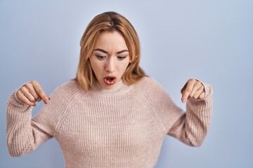 Wall Mural - Hispanic woman standing over blue background pointing down with fingers showing advertisement, surprised face and open mouth