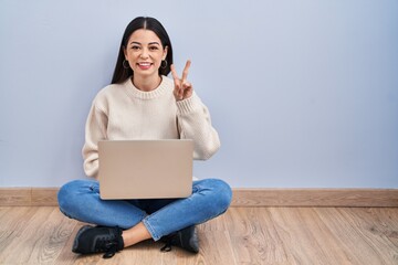 Sticker - Young woman using laptop sitting on the floor at home showing and pointing up with fingers number two while smiling confident and happy.