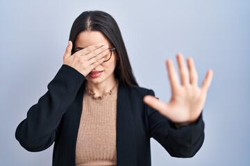 Canvas Print - Young brunette woman standing over blue background covering eyes with hands and doing stop gesture with sad and fear expression. embarrassed and negative concept.