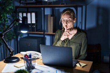 Wall Mural - Young blonde woman working at the office at night asking to be quiet with finger on lips. silence and secret concept.