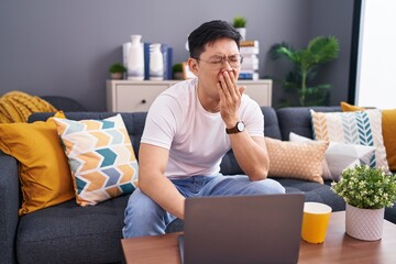 Wall Mural - Young asian man using laptop at home sitting on the sofa bored yawning tired covering mouth with hand. restless and sleepiness.