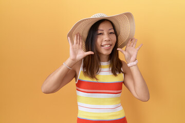 Wall Mural - Middle age chinese woman wearing summer hat over yellow background smiling cheerful playing peek a boo with hands showing face. surprised and exited