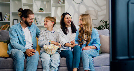 Attractive happy smiling parents and their two kids eating popcorn while sitting on the couch in the living room and watching TV. Cheerful mother, father, son and daughter spend time together at home.