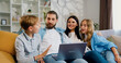 © serg - Good looking young family parents daughter and son using laptop while sitting in modern living room. Man, woman and children look at project of new house on computer and discuss something.