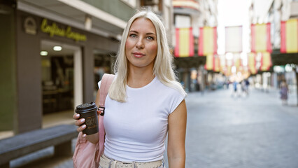 Poster - Relaxed and beautiful young blonde woman holding a hot take away coffee cup on a sunny city street, expressing her cool urban lifestyle outdoors.