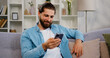 © serg - Hispanic Smiling man remote working online using smartphone while sitting on sofa couch in stylish cozy living room. Virtual home office, distance job. Freelancer Working.