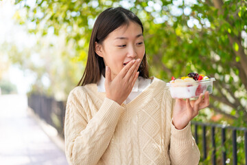 Wall Mural - Young Chinese woman holding a bowl of fruit at outdoors with surprise and shocked facial expression