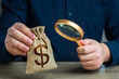 A man examines a dollar money bag through a magnifying glass. Investigating capital origins. Anti money laundering, tax evasion. Study terms and conditions on deposit or loan. Find investments