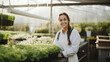 A cute lady with white apron standing and smiling in front of a green house nursery pots 