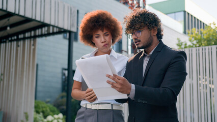 Two confused anxious diverse coworkers walking in city check papers bad result talking problem Indian businessman business talk with African businesswoman boss leader showing documents difficulties