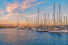 Yachts Moored In The Port Of Barcelona At Sunset, Spain. Many Boats With Masts In The Bay Of The Mediterranean Sea Against The Backdrop Of The City Coastline Illuminated By The Rays Of The Setting Sun