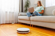 © spyrakot - Robotic vacuum cleaner cleaning room while woman resting on sofa using laptop