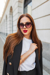 Wall Mural - Beautiful successful fashion business girl with vintage sunglasses with red lips in fashionable casual clothes with a black blazer walks in the city. Pretty chic woman